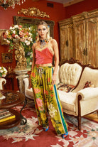 A person wearing olive floral pants with a striped pattern and side pockets, paired with a red top, standing in a room with classic furniture and floral decorations.